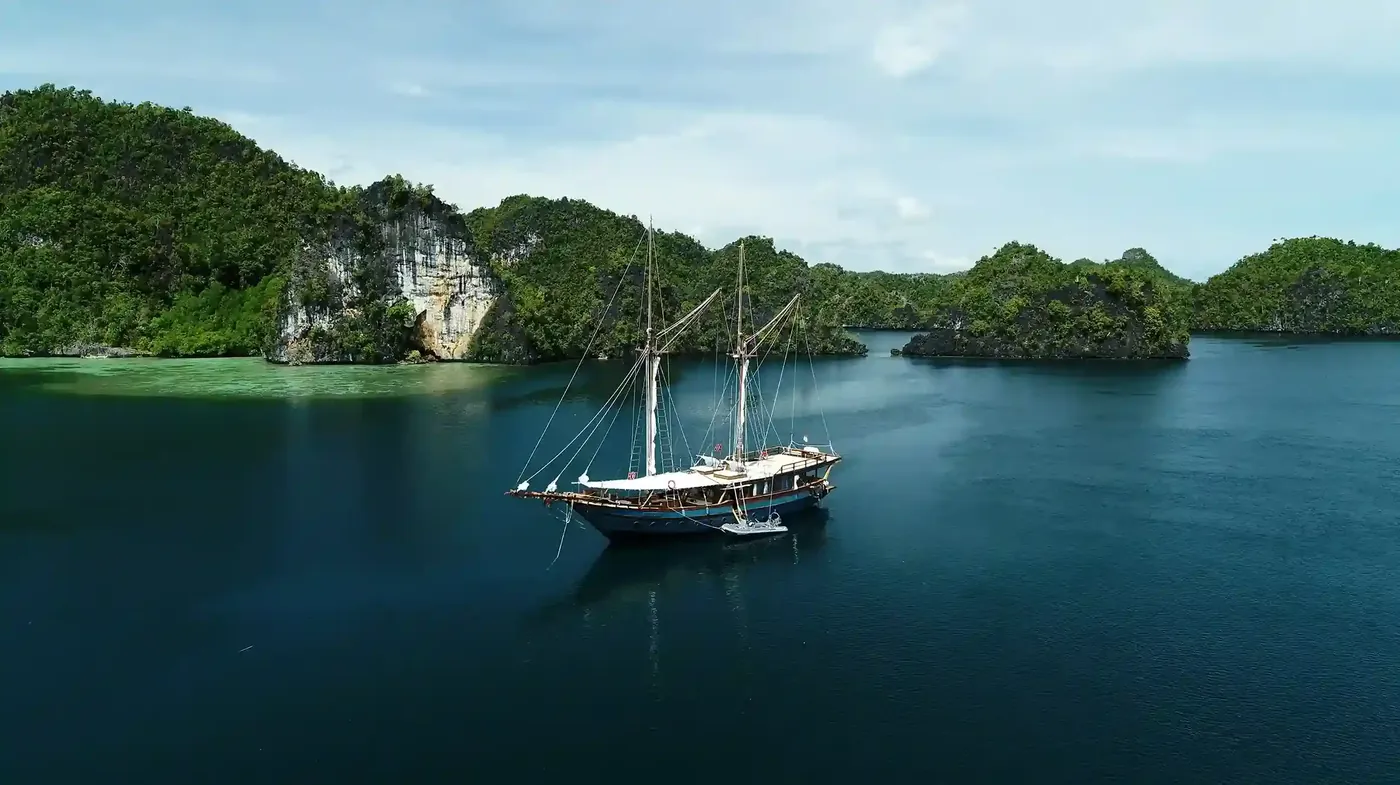 A masted phinisi lying at anchor in still water below the forested cliffs of Raja Ampat