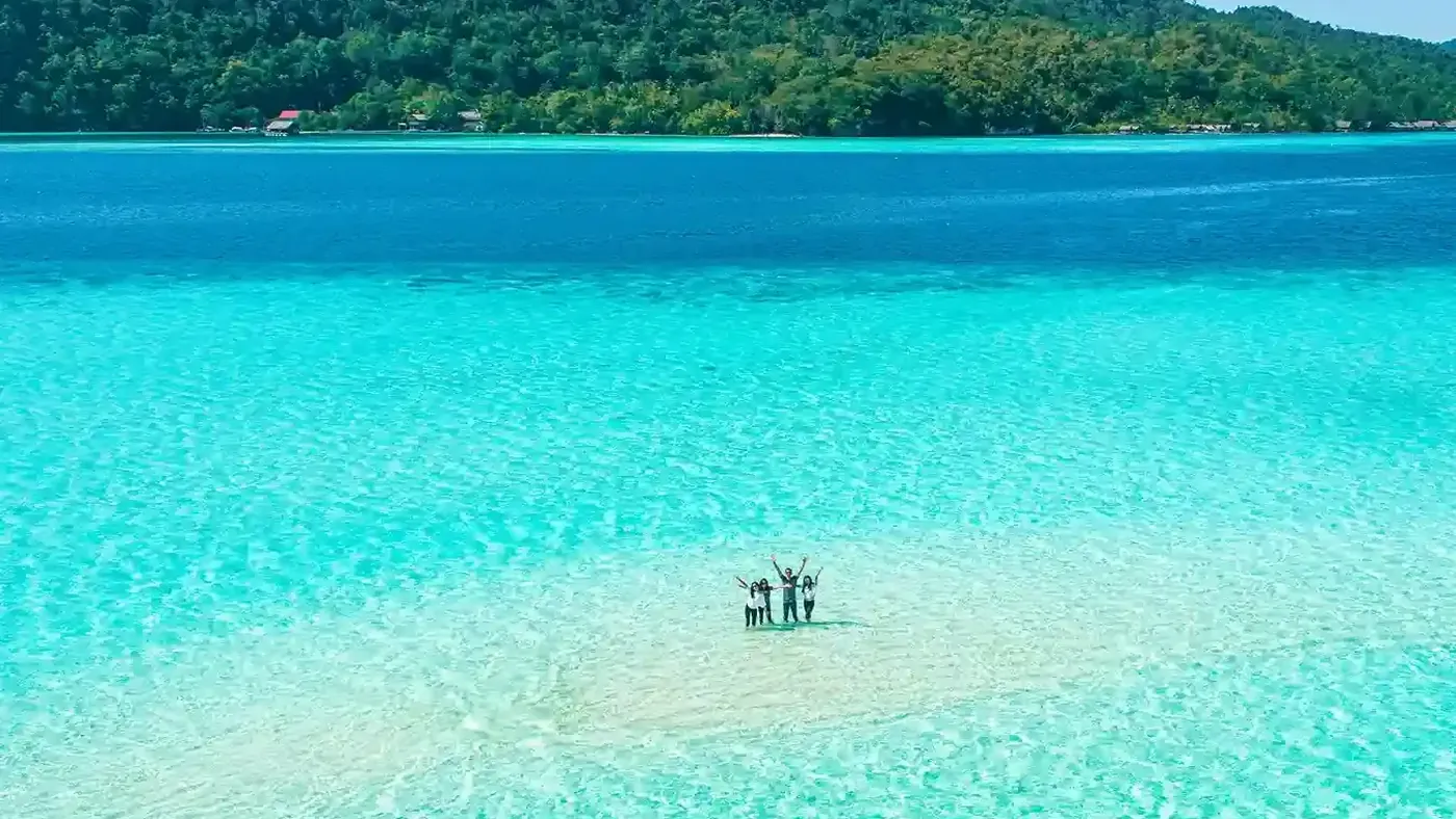 A pale sandbar surfacing in shallow turquoise water, with a jungle-covered island beyond